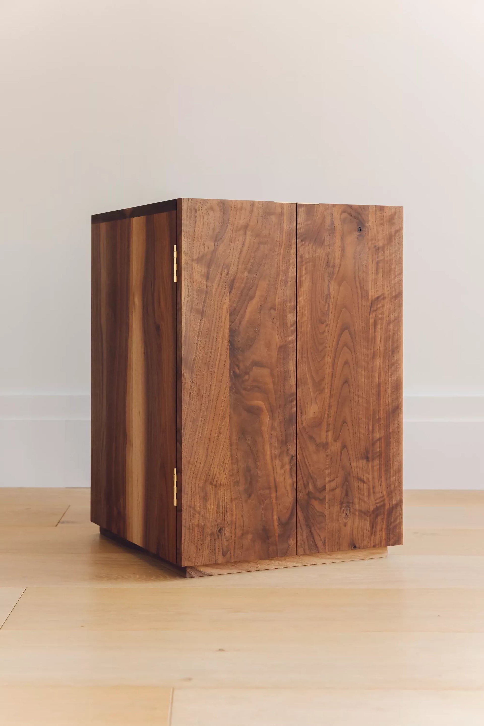 A wooden bedside cabinet with twin doors, an adjustable internal shelf, and solid brass butt hinges, shown in a natural light setting,  available  to buy on the-salon.com. SALON curates the finest , unique, handmade pieces from the interior industry's best makers and brands. Shop luxury furniture, lighting, rugs, art, antiques and vintage pieces and accessories from SALON interior designed rooms.