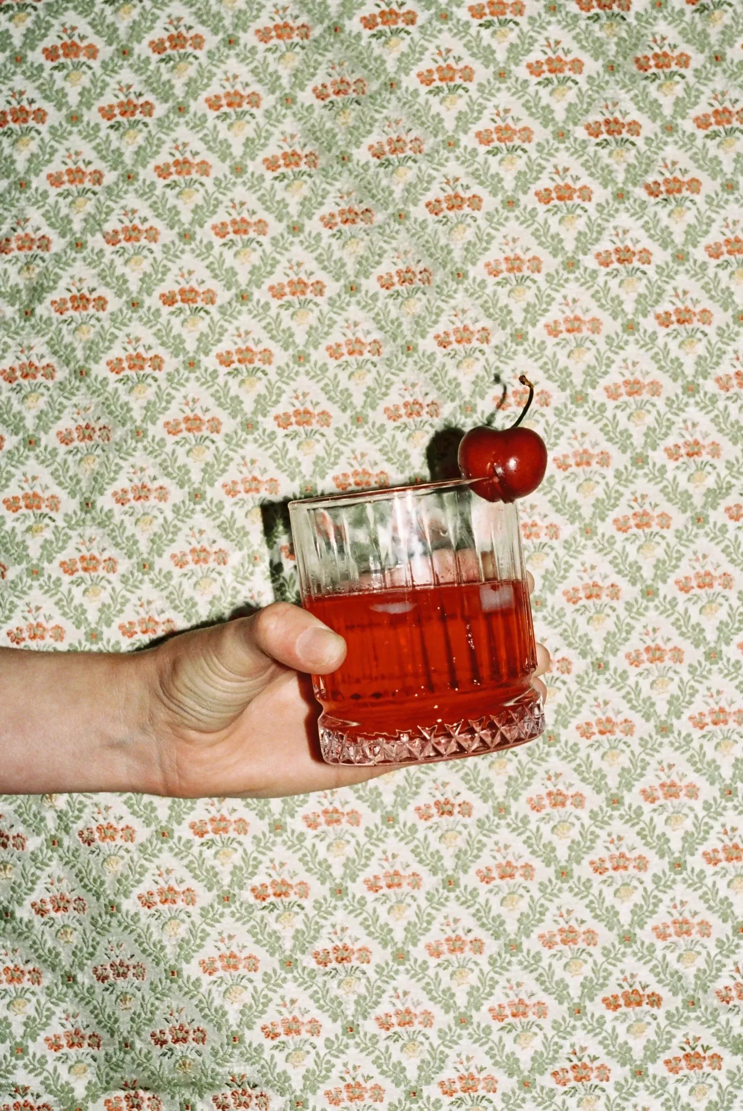 A textile fabric with a jacquard weave pattern featuring vibrant red poppies in a scallop shell vase, surrounded by foliage arranged in a trellis style, in the background of a person holding a red drink in a glass with a cherry on top, available  to buy on the-salon.com. SALON curates the finest , unique, handmade pieces from the interior industry's best makers and brands. Shop luxury furniture, lighting, rugs, art, antiques and vintage pieces and accessories from SALON interior designed rooms.