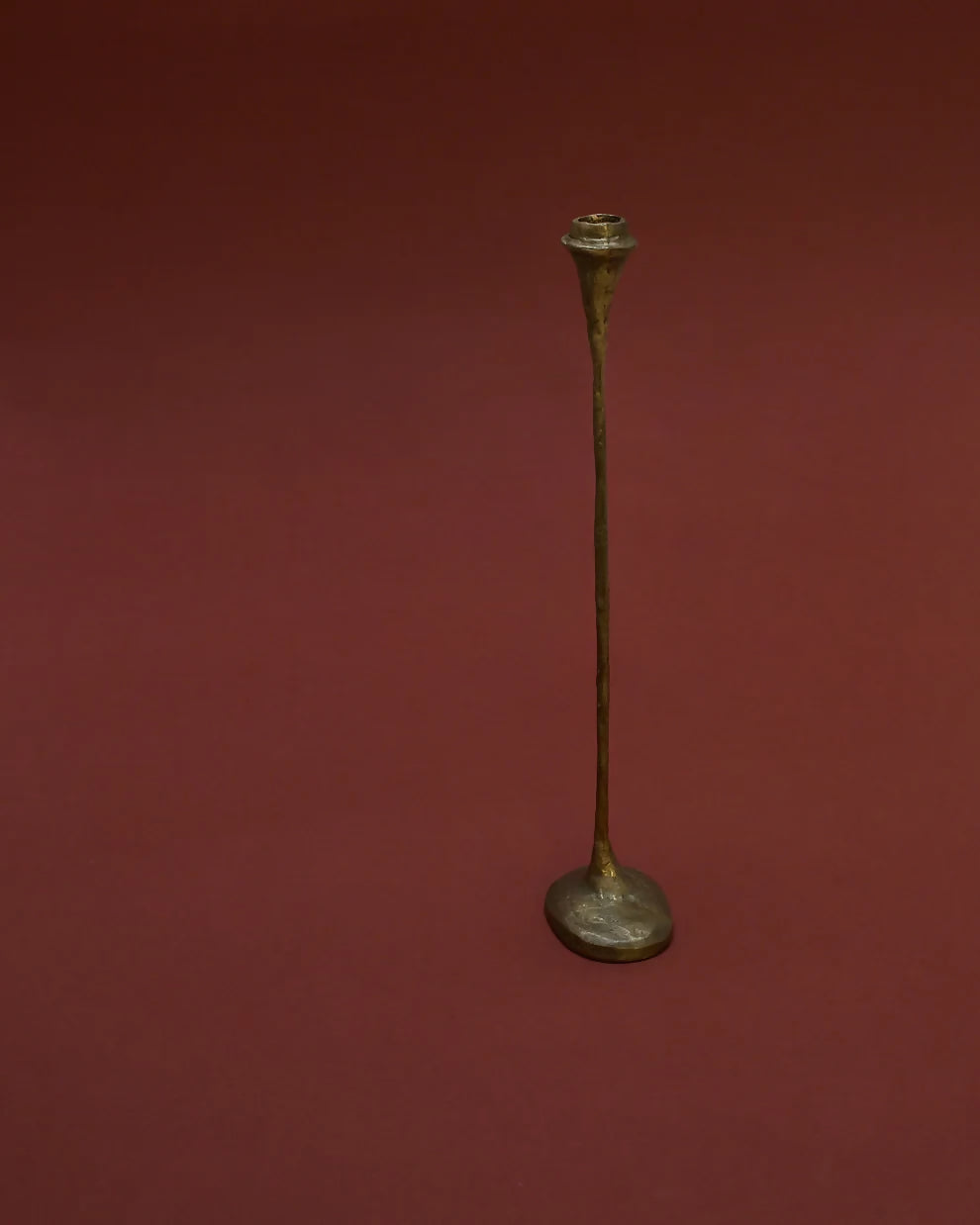 A tall, solid bronze candle holder with a textured finish, standing on a flat surface with a red background,  available  to buy on the-salon.com. SALON curates the finest , unique, handmade pieces from the interior industry's best makers and brands. Shop luxury furniture, lighting, rugs, art, antiques and vintage pieces and accessories from SALON interior designed rooms.