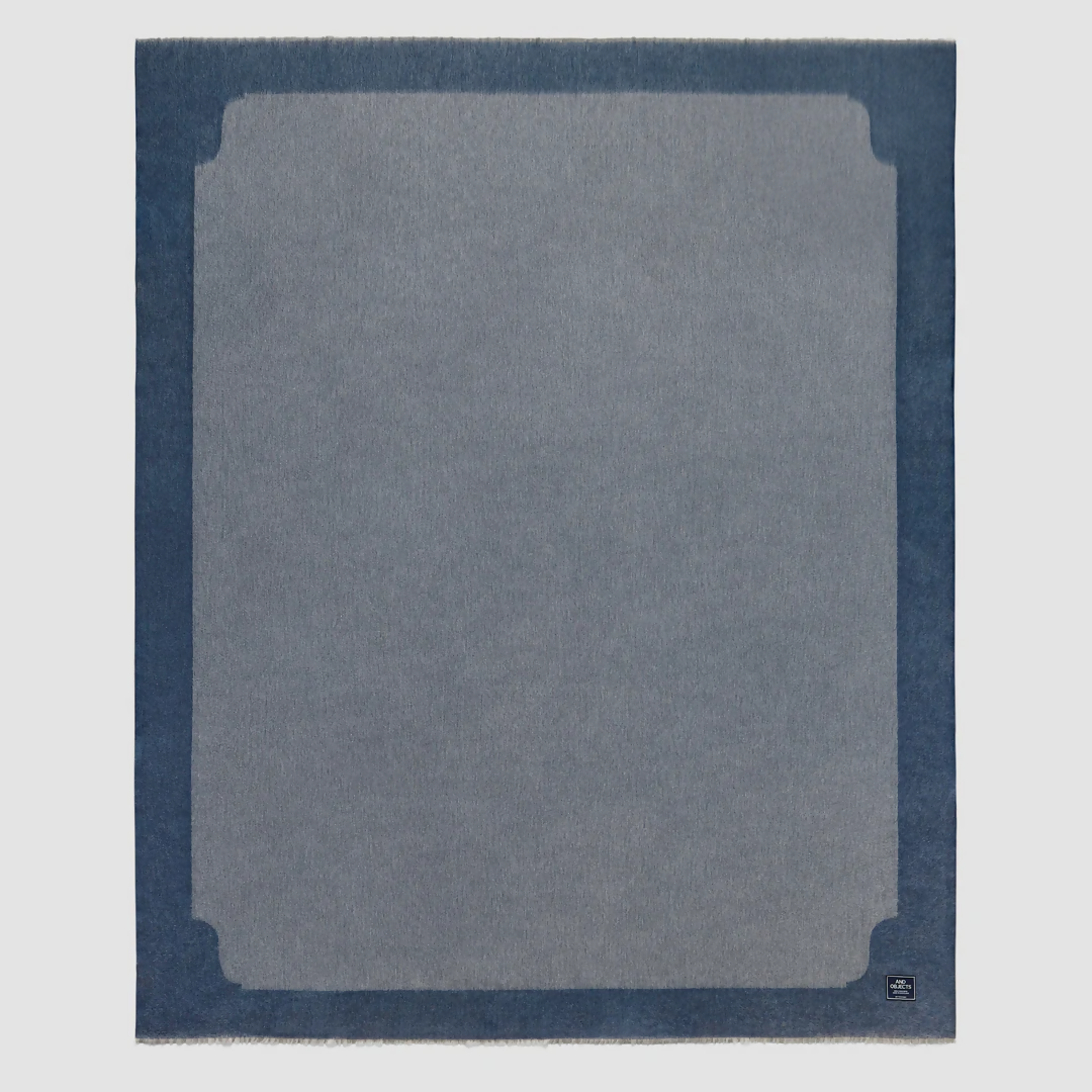 A folded slate blue cashmere throw blanket with a decorative border detail,  available  to buy on the-salon.com. SALON curates the finest , unique, handmade pieces from the interior industry's best makers and brands. Shop luxury furniture, lighting, rugs, art, antiques and vintage pieces and accessories from SALON interior designed rooms.