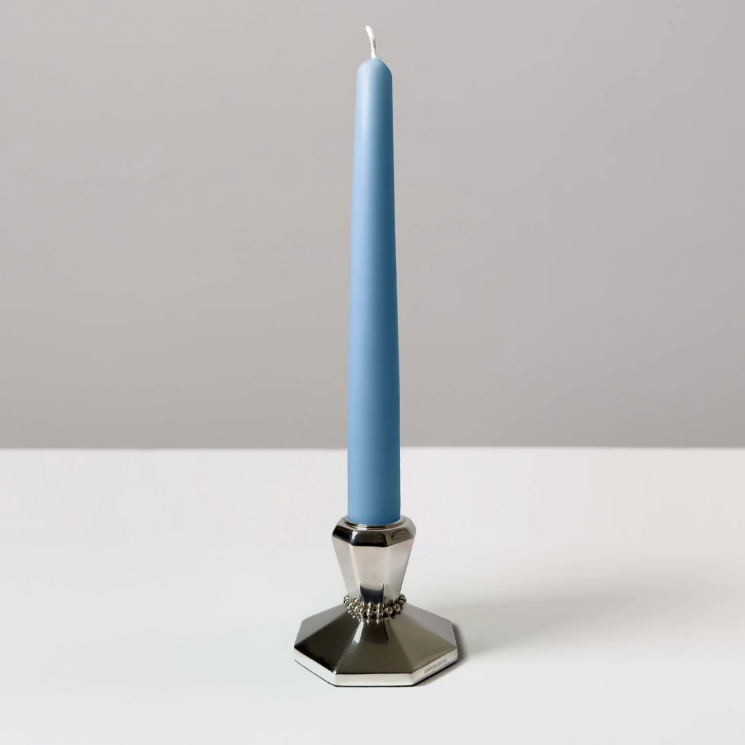 A silver candle stick with a blue candle on a white background, featuring intricate beaded detailing around the neck and an angular base,  available  to buy on the-salon.com. SALON curates the finest , unique, handmade pieces from the interior industry's best makers and brands. Shop luxury furniture, lighting, rugs, art, antiques and vintage pieces and accessories from SALON interior designed rooms.