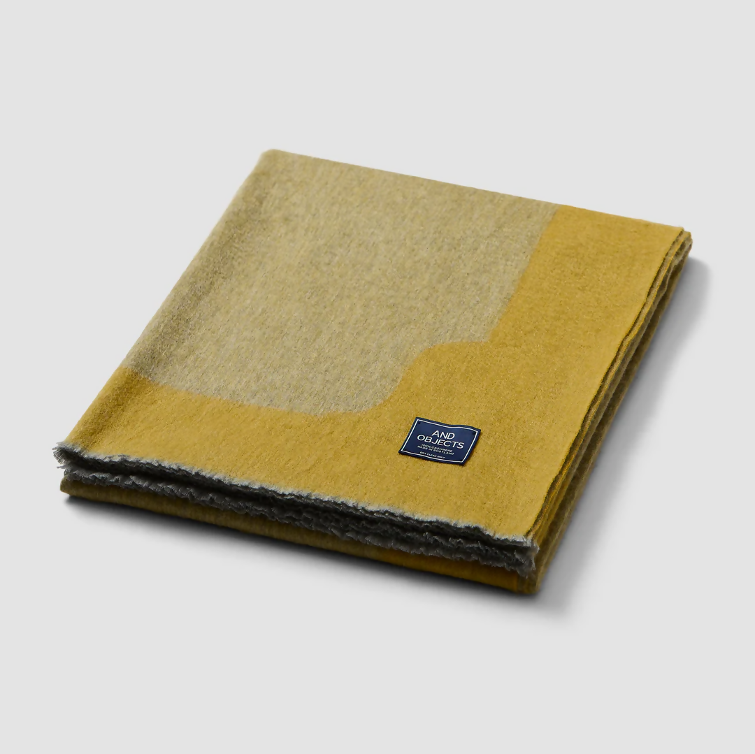 A folded gold-colored cashmere throw blanket with a decorative border, displaying a small blue and silver brand tag on the bottom right corner,  available  to buy on the-salon.com. SALON curates the finest , unique, handmade pieces from the interior industry's best makers and brands. Shop luxury furniture, lighting, rugs, art, antiques and vintage pieces and accessories from SALON interior designed rooms.
