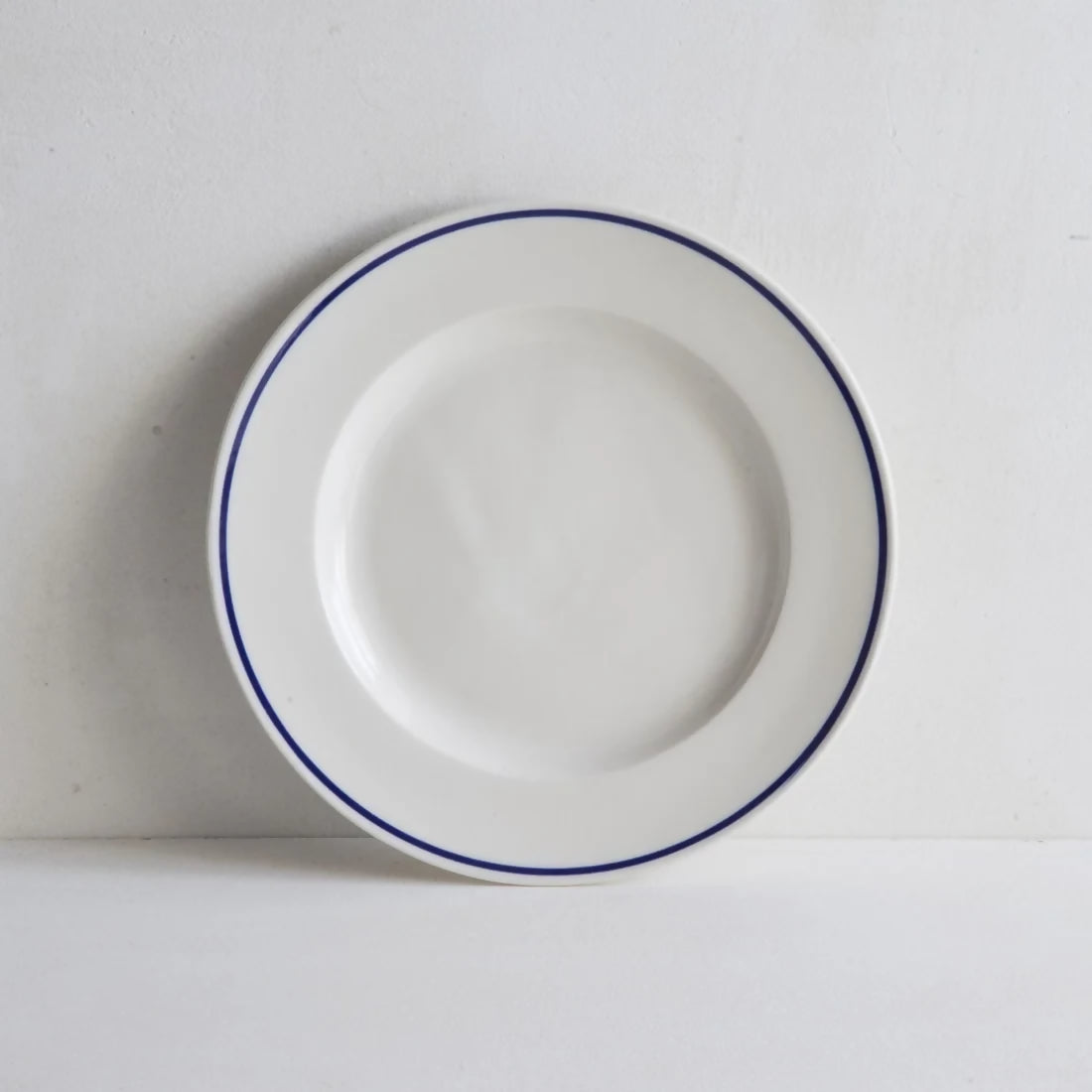 A round white porcelain side plate with a rich cobalt blue line along the rim, displayed against a white background, available  to buy on the-salon.com. SALON curates the finest , unique, handmade pieces from the interior industry's best makers and brands. Shop luxury furniture, lighting, rugs, art, antiques and vintage pieces and accessories from SALON interior designed rooms