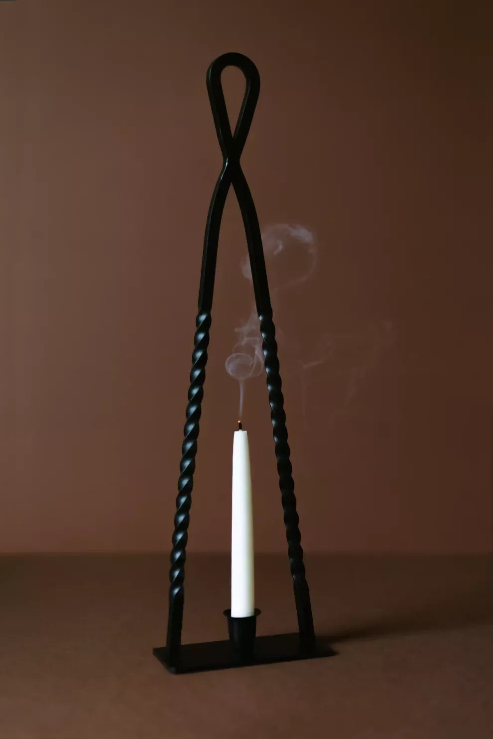 A black, handmade steel Alchemy candle holder by HUEY, displayed on a brown background with a candle inside it, surrounded by cherries on the table, available  to buy on the-salon.com. SALON curates the finest , unique, handmade pieces from the interior industry's best makers and brands. Shop luxury furniture, lighting, rugs, art, antiques and vintage pieces and accessories from SALON interior designed rooms.